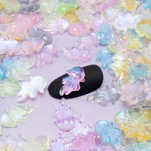 May include: Assortment of iridescent pastel-colored plastic jellyfish charms. The charms are scattered on a purple surface with a black nail file in the center. The nail file has a single iridescent jellyfish charm on top.
