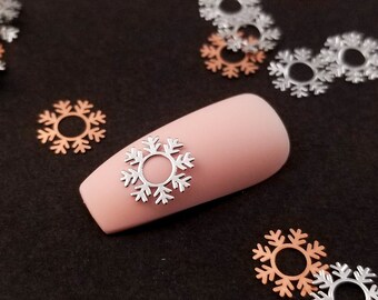 May include: A close-up of a fingernail with a silver snowflake nail art design. The nail is surrounded by a variety of silver and copper snowflake nail charms.
