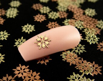 May include: A pink nail with a gold snowflake design on top of a black surface covered in gold and copper snowflake confetti.
