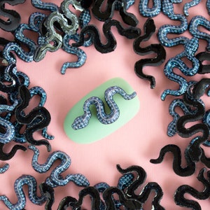 May include: A close-up of a light blue nail with a black and blue snake design. The nail is surrounded by many black and blue snake-shaped rhinestones on a pink background.
