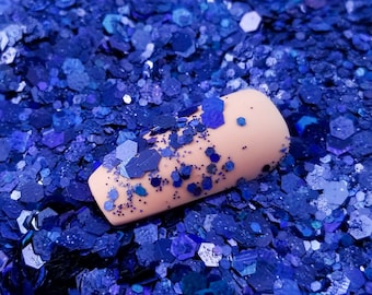 May include: A close-up of a light pink nail with blue glitter on it. The nail is surrounded by a pile of loose blue glitter.