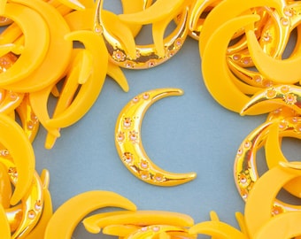 May include: A close-up of a group of yellow crescent moon-shaped charms. The charms are made of plastic and have a shiny, metallic finish. Some of the charms have small, clear rhinestones embedded in the surface.
