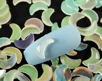 May include: A close-up of iridescent holographic crescent moon shaped nail art embellishments. The embellishments are scattered on a black surface. A light blue nail with a crescent moon embellishment is in the center of the image.