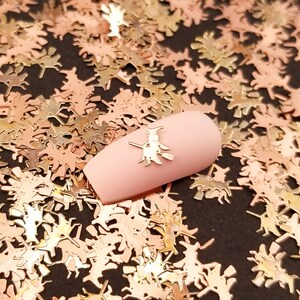 May include: A close-up of a pink nail with a gold witch charm on top. The nail is surrounded by a scattering of gold glitter in the shape of witches on broomsticks.