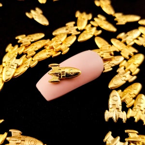 May include: Gold-colored rocket ship nail charms scattered on a black surface. One rocket ship charm is on a pink fingernail.