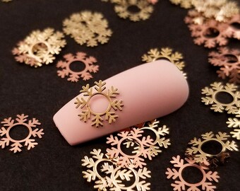 May include: Gold and rose gold snowflake nail charms scattered on a black surface. One snowflake charm is placed on a pink nail.