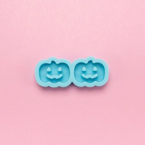 May include: Two light blue silicone molds shaped like pumpkins with jack-o'-lantern faces. The molds are on a pink background.
