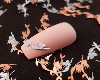 May include: A close-up of a light pink nail with a silver mermaid cutout on top. The nail is surrounded by a scattering of silver and copper mermaid cutouts on a black background.