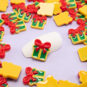 May include: Assortment of small, colorful, plastic Christmas gift box charms in red, green, and yellow. The charms are scattered on a purple surface.
