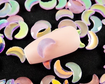 May include: Iridescent crescent moon-shaped nail art decorations scattered on a black surface. The sequins have a shimmering, multi-colored effect, reflecting hues of purple, green, and yellow. A nail with a moon design is also present.