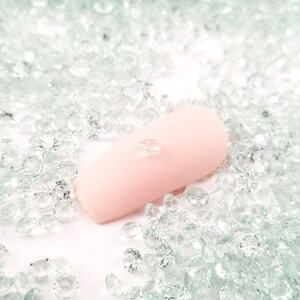 May include: A light pink nail with a single clear rhinestone applied. The nail is surrounded by a multitude of small, clear rhinestones scattered across the surface, creating a sparkling effect. The overall theme is nail art.
