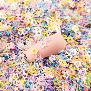 May include: A close-up of a pile of colorful star-shaped glitter. The glitter is a mix of pastel pink, blue, yellow, and green. The glitter is perfect for adding a touch of sparkle to crafts, nail art, and other projects.