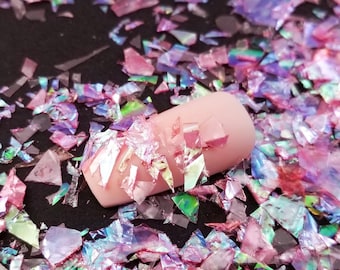 Puede incluir: Primer plano de escamas de purpurina iridiscente rosa y púrpura sobre una superficie negra. Se muestra una uña con la purpurina aplicada.