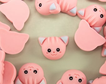 May include: A collection of pink cat-shaped resin charms with white ears and black eyes. The charms are scattered on a light beige surface.