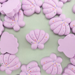 May include: A collection of light purple seashell-shaped embellishments with colorful dot accents. The image shows various sizes and styles of the decorative pieces, perfect for crafting or embellishing projects. The background is a light green color.