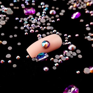 May include: A close-up of a fingernail with a single iridescent rhinestone on top. The nail is surrounded by a scattering of iridescent rhinestones of various sizes and shapes on a black background.