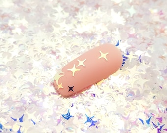 May include: A single pink nail with gold star accents on a background of iridescent white star-shaped glitter.