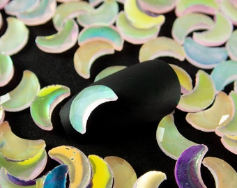 May include: A close-up of iridescent crescent moon shaped nail charms. The charms are scattered on a black surface.
