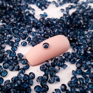 May include: A close-up of a single blue rhinestone on a fingernail surrounded by a scattering of smaller blue rhinestones.