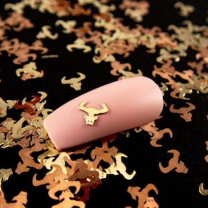 May include: A pink nail with a gold bull charm on a black background surrounded by gold and copper metallic confetti.