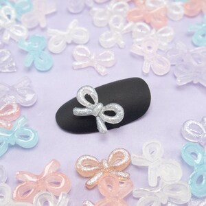 May include: A close-up of a black nail with a silver glitter bow on top. The nail is surrounded by a variety of colorful glitter bows.