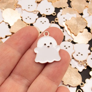 May include: A hand holding a white metal ghost charm with a gold loop. The charm has a simple, smiling face. The background is a pile of similar charms, some gold and some white.