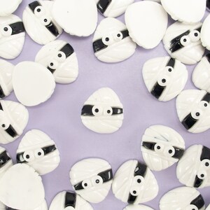 May include: A collection of white and black mummy-themed embellishments with googly eyes. The embellishments are scattered on a light purple surface.