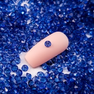 May include: A close-up of a light pink nail with a single blue rhinestone on top. The nail is surrounded by a scattering of tiny blue rhinestones.