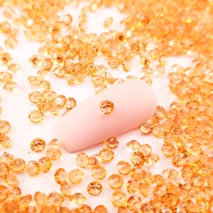 May include: A close-up of a pink fingernail with a single clear rhinestone on top. The fingernail is surrounded by a scattering of small, orange-colored rhinestones.