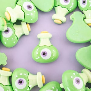 May include: A collection of green and purple resin bottle charms with a single eye design. The charms are shaped like bottles and have a round eye with a purple pupil. The charms are scattered on a light purple background.