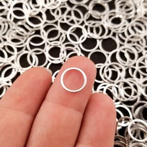 May include: A close-up of a silver jump ring held between two fingers. The jump ring is a small, circular piece of metal with a gap in it. The jump ring is surrounded by a large quantity of similar jump rings.