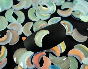 May include: A close-up of iridescent, light blue crescent moon shaped nail charms. The charms are scattered on a black surface. One charm is placed on a black nail.