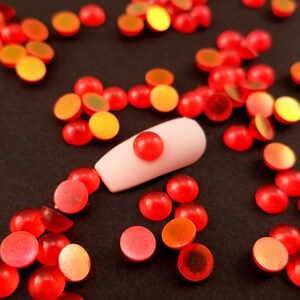 May include: A collection of small, red, half-circle shaped beads with a glossy finish. Some beads have a gradient of orange and yellow. A light pink nail is in the center of the image, with a single red bead on top. The beads are scattered on a dark surface.