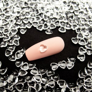 May include: A pink nail with a clear heart-shaped rhinestone on top. The nail is surrounded by clear heart-shaped confetti.