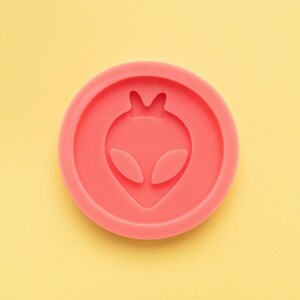 May include: A pink silicone mold with an alien head design. The mold is circular and has a smooth surface.