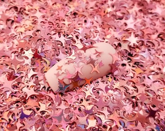 May include: A close-up of a pink nail with iridescent pink star-shaped glitter on top. The nail is surrounded by a bed of loose iridescent pink star-shaped glitter.