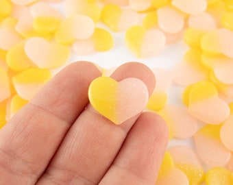 May include: A hand holding a single heart-shaped candy with a yellow top and a pink bottom. The candy is surrounded by a large quantity of similar candies in a mix of yellow and pink.