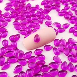May include: A close-up of a pink fingernail with a single purple teardrop-shaped rhinestone on top. The fingernail is surrounded by a scattering of many small purple teardrop-shaped rhinestones.