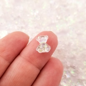 May include: A small, clear iridescent resin bear charm. The bear is sitting with its paws in front of it. The charm is perfect for adding a touch of whimsy to jewelry, crafts, and other projects.