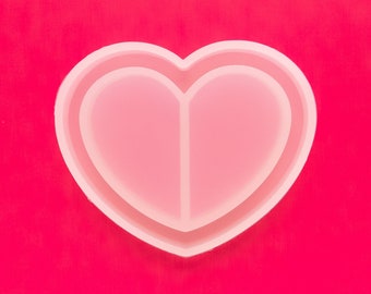 Puede incluir: Un molde de silicona blanco en forma de corazón con dos compartimentos separados. El molde está sobre un fondo rosa brillante.