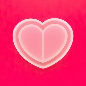 May include: A white silicone heart-shaped mold with two separate compartments. The mold is on a bright pink background.