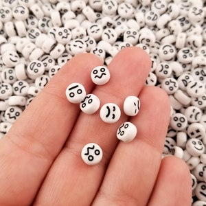 May include: A hand holding white plastic beads with different black face expressions. The beads are round and have a hole in the center for stringing.