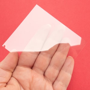 May include: A clear plastic sheet with a slightly frosted appearance. The sheet is being held in a hand.
