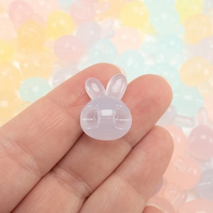 May include: A translucent lavender bunny-shaped charm with two upright ears and a rounded body. The charm has a small clip on the back. The background is filled with pastel-colored, round beads.