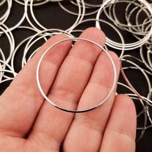 May include: A silver metal ring, approximately 2 inches in diameter, is held in a person's hand. The ring is smooth and has a polished finish. The ring is part of a large pile of similar rings.