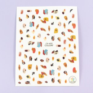 May include: A sheet of colourful nail art stickers with abstract shapes in shades of brown, orange, yellow, blue, and grey. The sheet is labelled "CA-603 COLORF".