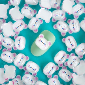 May include: A close-up of a group of white bear-shaped charms with pink ears and blue eyes. The charms are scattered on a turquoise surface with a single charm on a light green surface.