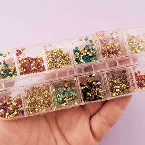 May include: A clear plastic box filled with 12 small compartments, each containing a different colour of rhinestones. The rhinestones are small and sparkly, and they are arranged in a neat and organised fashion.