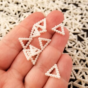May include: A hand holding a handful of small white pearl triangle shapes. The triangles are made of small white pearls strung together in a triangular shape.
