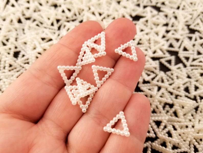 May include: A hand holding a handful of small white pearl triangle shapes. The triangles are made of small white pearls strung together in a triangular shape.